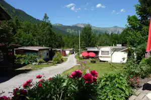 Campingplatz mit gepflastertem Weg, Wohnwagen und roten Markisen, Blumenbeeten und Blick auf bewaldete Berge.