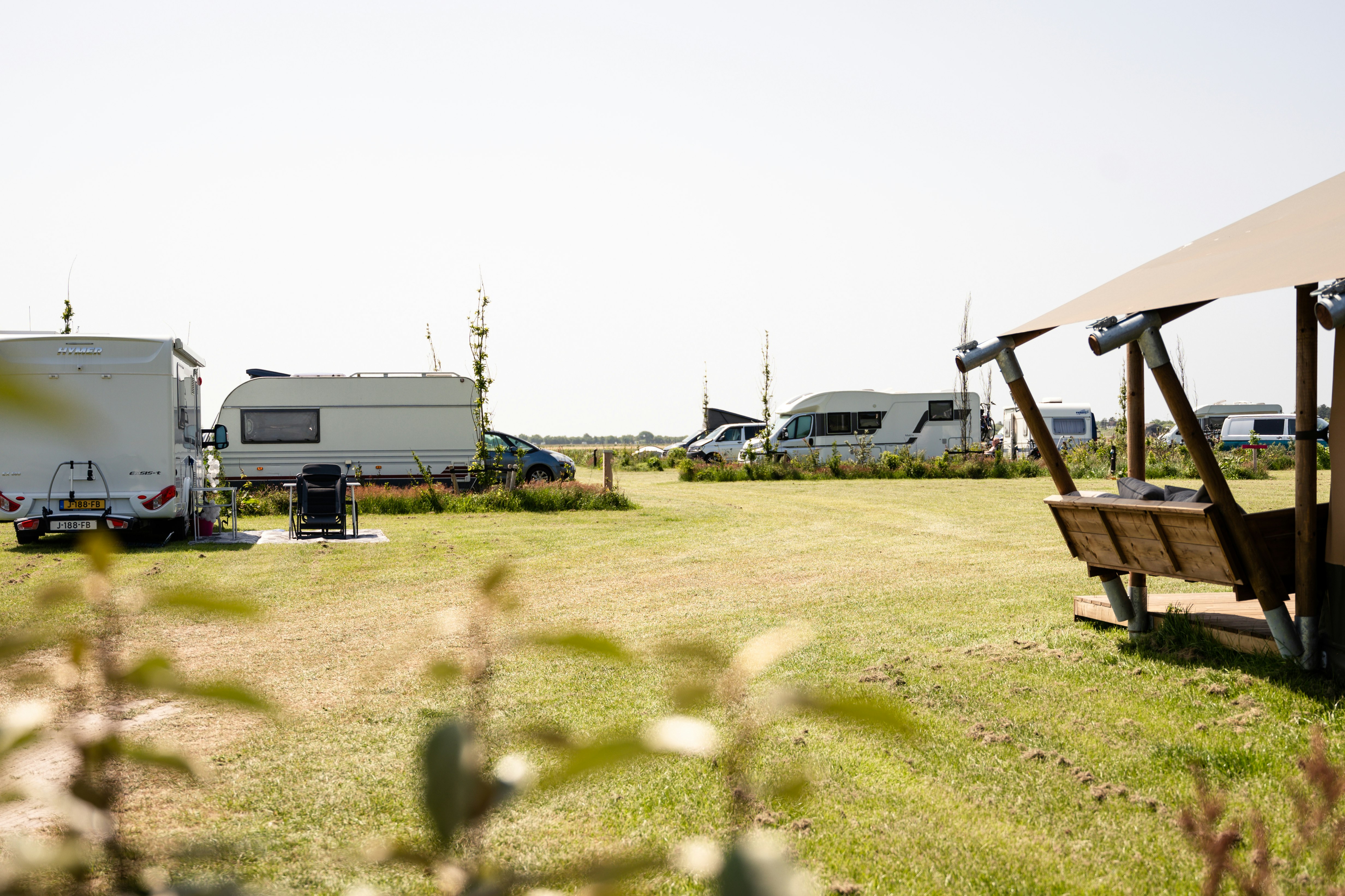 Rasenfläche auf dem Campingplatz mit Sitzbank unter einem Sonnendach, im Hintergrund Wohnwagen und weitere Stellplätze.