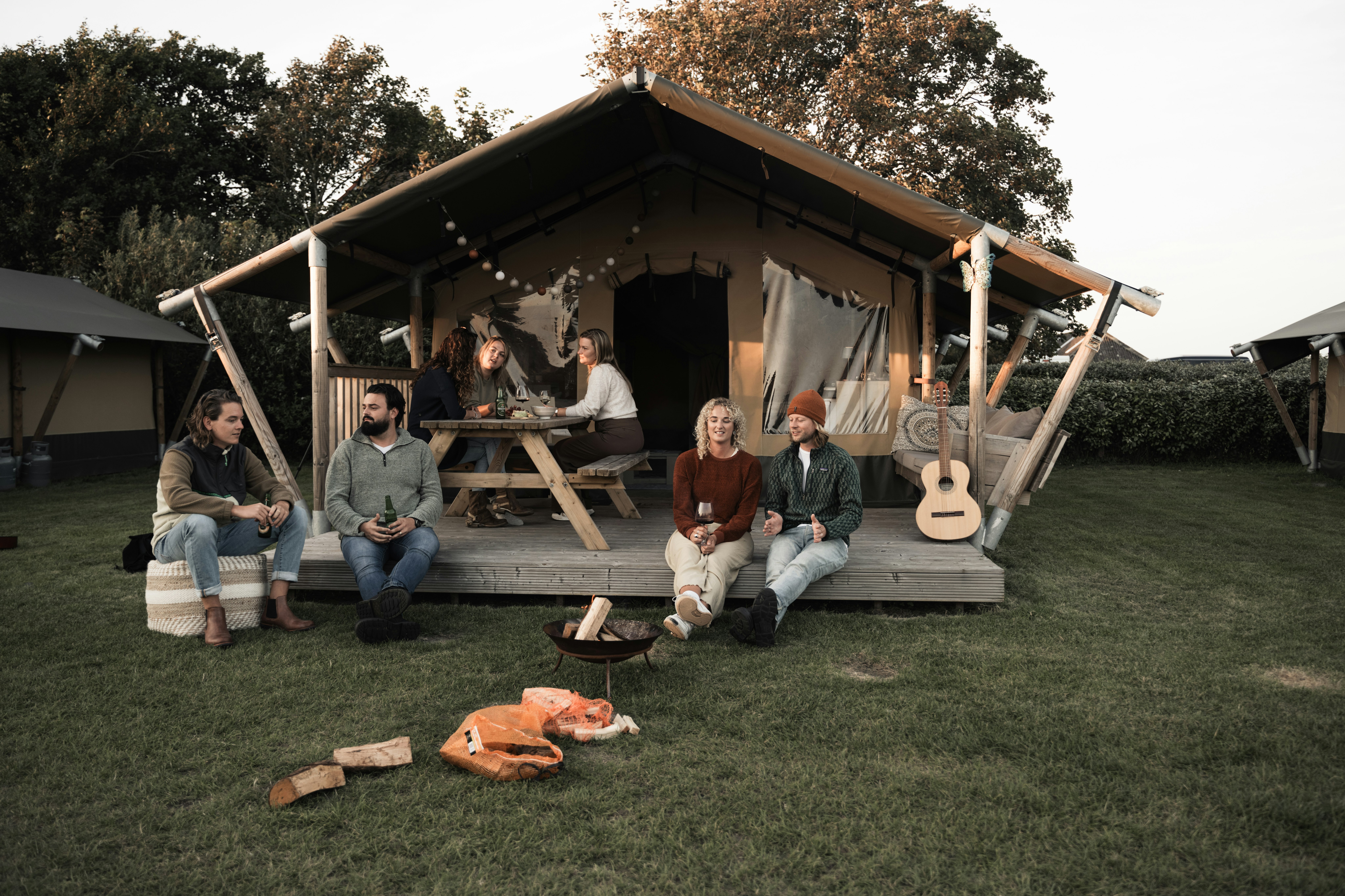 Campinggruppe vor einem Holz-Zelt mit Veranda, Sitzbank und Tisch; Gitarren an der Wand, Rasenfläche im Hintergrund.