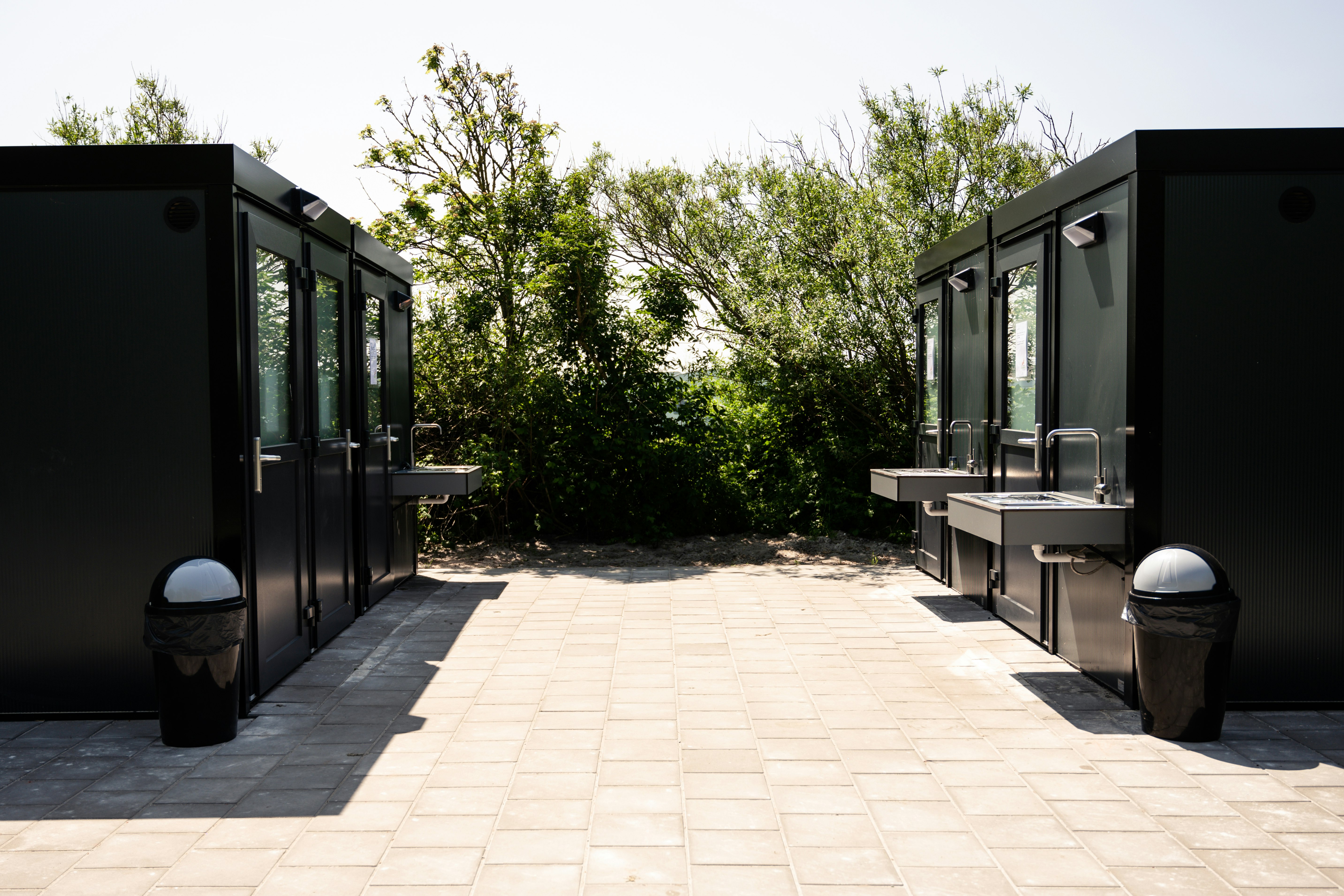 Gepflasterter Weg zwischen zwei modernen Camping-Modulen mit Türen, daneben Mülltonnen und Hecken im Hintergrund.