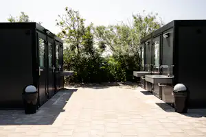 Gepflasterter Weg zwischen zwei modernen Camping-Modulen mit Türen, daneben Mülltonnen und Hecken im Hintergrund.