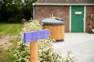 Hinweisschild mit handschriftlicher Aufschrift auf einem Holzpfosten vor einem Stellplatz mit Mülltonnen und Gebäude.