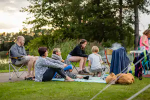 Familie sitzt auf Campingstühlen auf einer Wiese, Decke und Rucksack daneben, im Hintergrund Bäume und ein Wohnwagen.