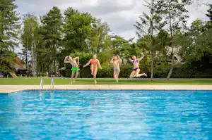 Kinder springen am Camping-Schwimmbecken, im Hintergrund Bäume und ein grüner Rasenbereich.