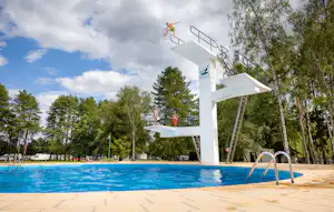 Sprungturm am Camping-Pool mit Leiter und Plattform, umgeben von Bäumen und blauem Himmel.