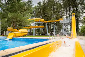 Gelbe Wasserrutsche am Campingplatz, Spritzwasser ins Becken, daneben Spielbereich mit Bäumen und Sonnenschirm.