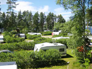 Wohnwagen auf einem Campingplatz zwischen Hecken und Bäumen, im Hintergrund weitere Fahrzeuge und Stellplätze.