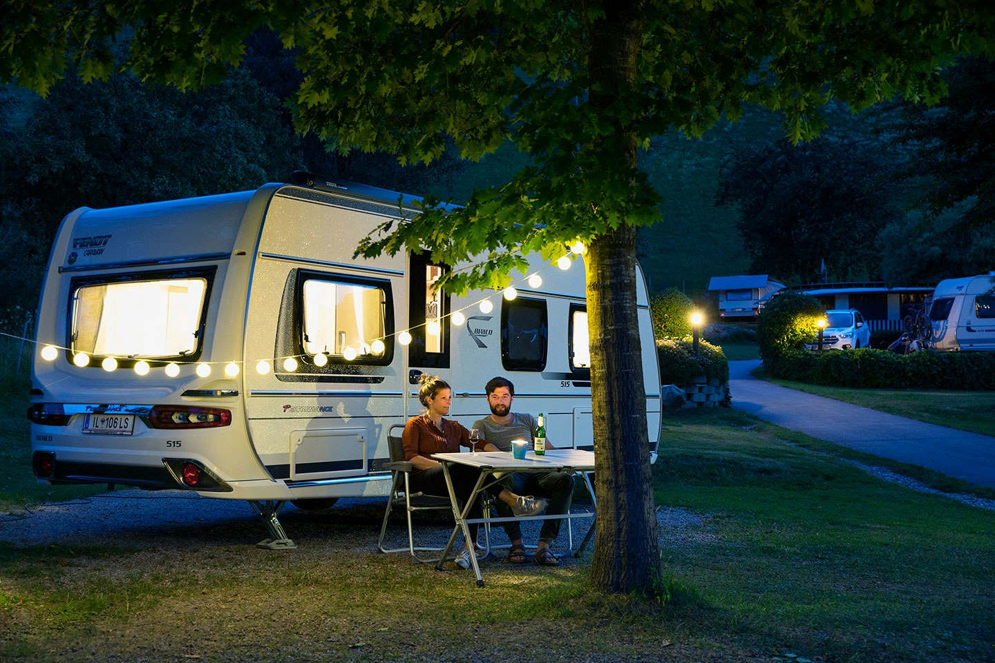 Wohnwagen auf dem Campingplatz mit Tisch und Stühlen vor dem Eingang, im Hintergrund weitere Fahrzeuge und Bäume.