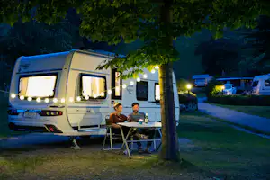 Wohnwagen auf dem Campingplatz mit Tisch und Stühlen vor dem Eingang, im Hintergrund weitere Fahrzeuge und Bäume.