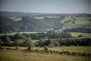 Blick auf eine hügelige Landschaft mit Radfahrern auf einem Feldweg, umgeben von Wiesen, Bäumen und entfernten Dörfern.