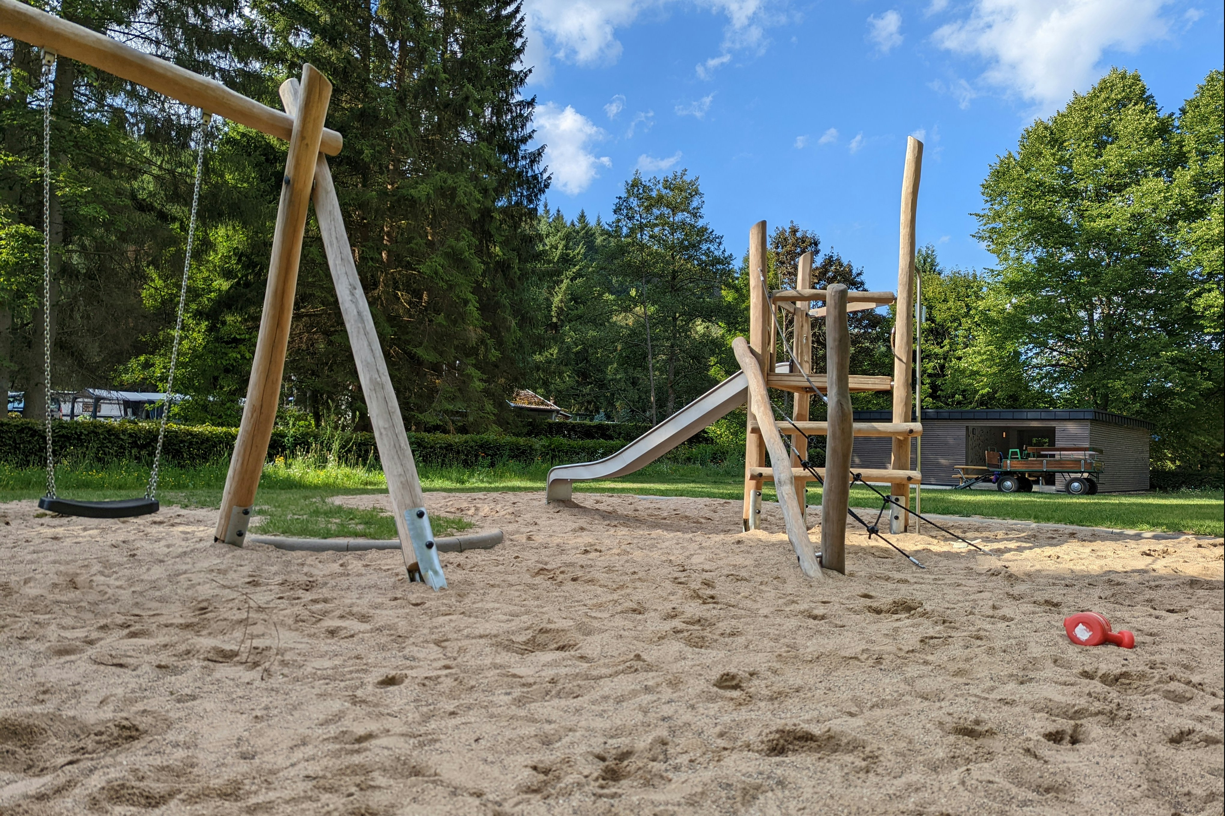 Spielplatz am Campingplatz mit Sandboden, Rutsche und Schaukel vor hohen Bäumen im Grünen.