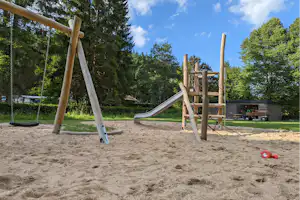 Spielplatz am Campingplatz mit Sandboden, Rutsche und Schaukel vor hohen Bäumen im Grünen.
