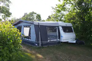 Wohnwagen mit Vorzelt auf einem Campingplatz, umgeben von Büschen und Bäumen, auf einer Rasenfläche.