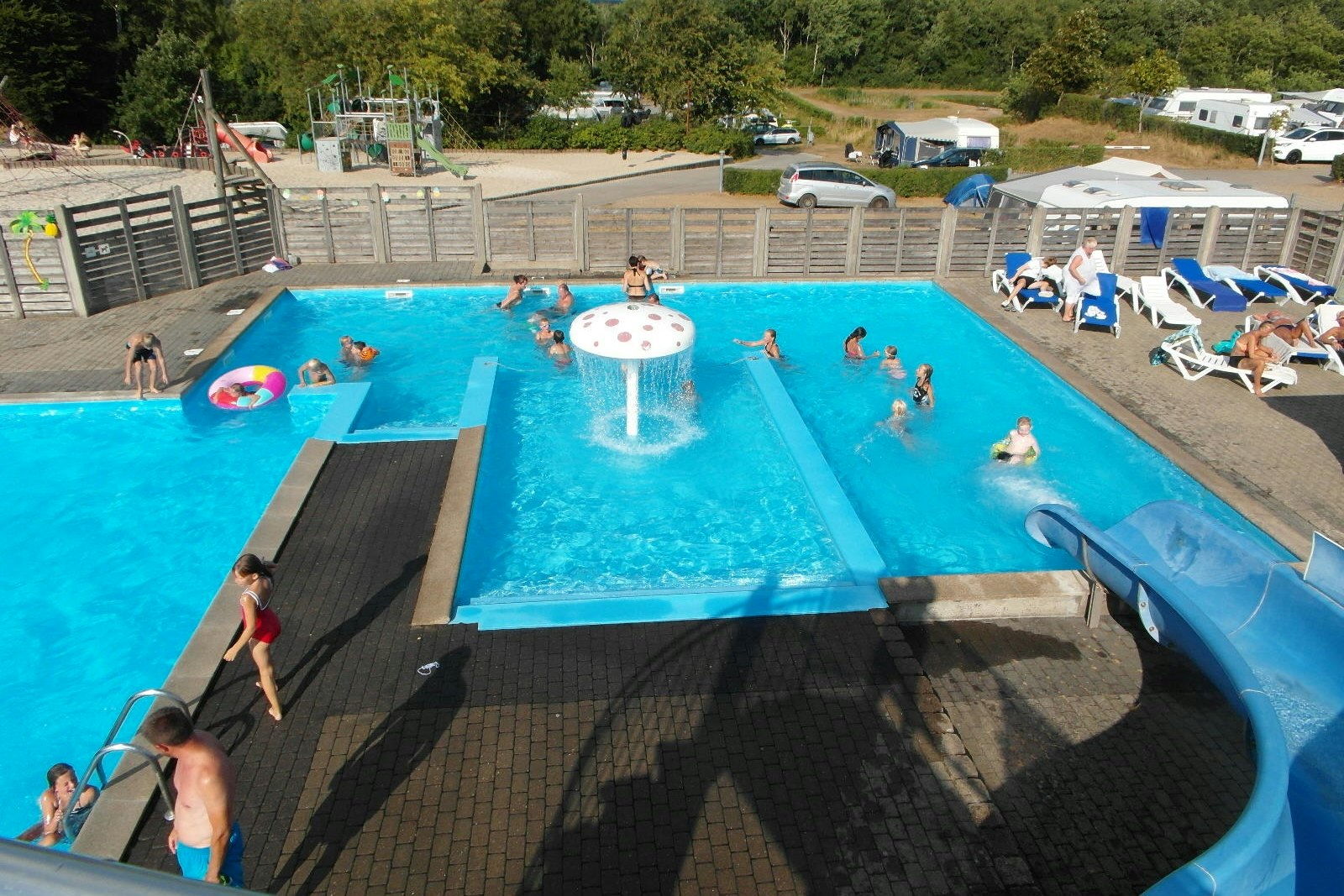 Camping Hessellund Sø - Swimmingpool auf dem Campingplatz mit Liegestühlen und Sonnenschirmen, im Hintergrund der Kinderspielplatz