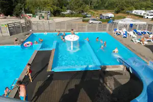 Camping Hessellund Sø - Swimmingpool auf dem Campingplatz mit Liegestühlen und Sonnenschirmen, im Hintergrund der Kinderspielplatz