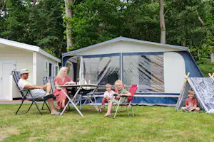 Camping mit Wohnwagen und Zelt, davor Stühle und Tisch auf einer Rasenfläche, im Hintergrund Bäume.