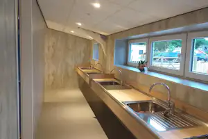 Camping-Küchenzeile mit Spüle, Wasserhahn und Arbeitsplatte, daneben Fenster mit Blick auf Gebäude und Sitzbereich.