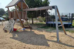 Spielplatz auf dem Campingplatz: Sandkasten, Klettergerüst und Trampolin neben Bäumen und einem Zaun.