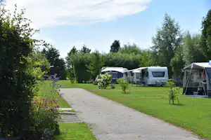 Campingplatz mit Wohnwagen und Zelt auf Stellplätzen, gepflasterter Weg, Rasen, Blumenbeete und Bäume im Hintergrund.