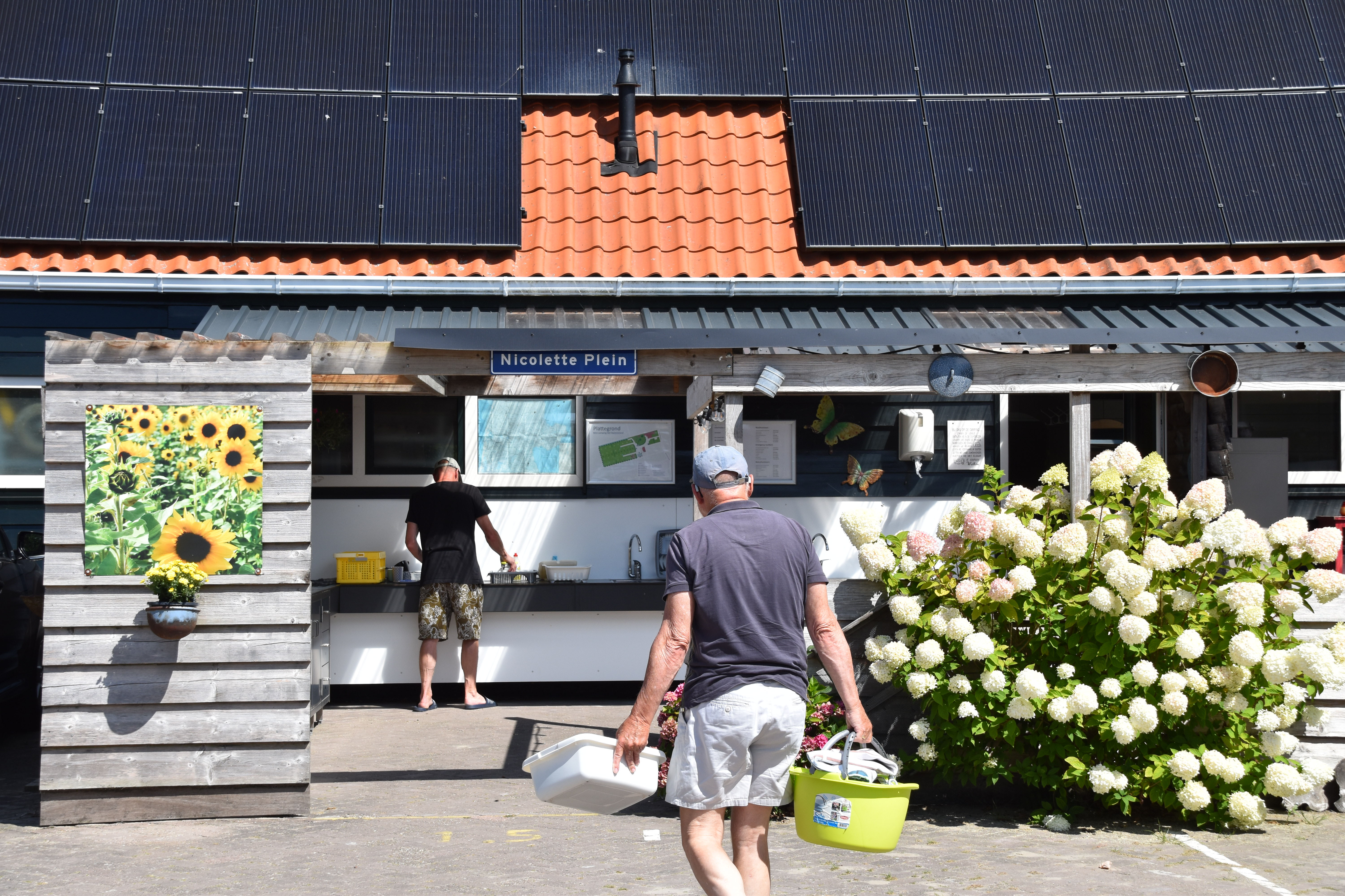 Rezeption eines Campingplatzes mit Blumenstrauß, Sonnenschirmen und Solarpaneln auf dem Dach, davor gehen zwei Personen.