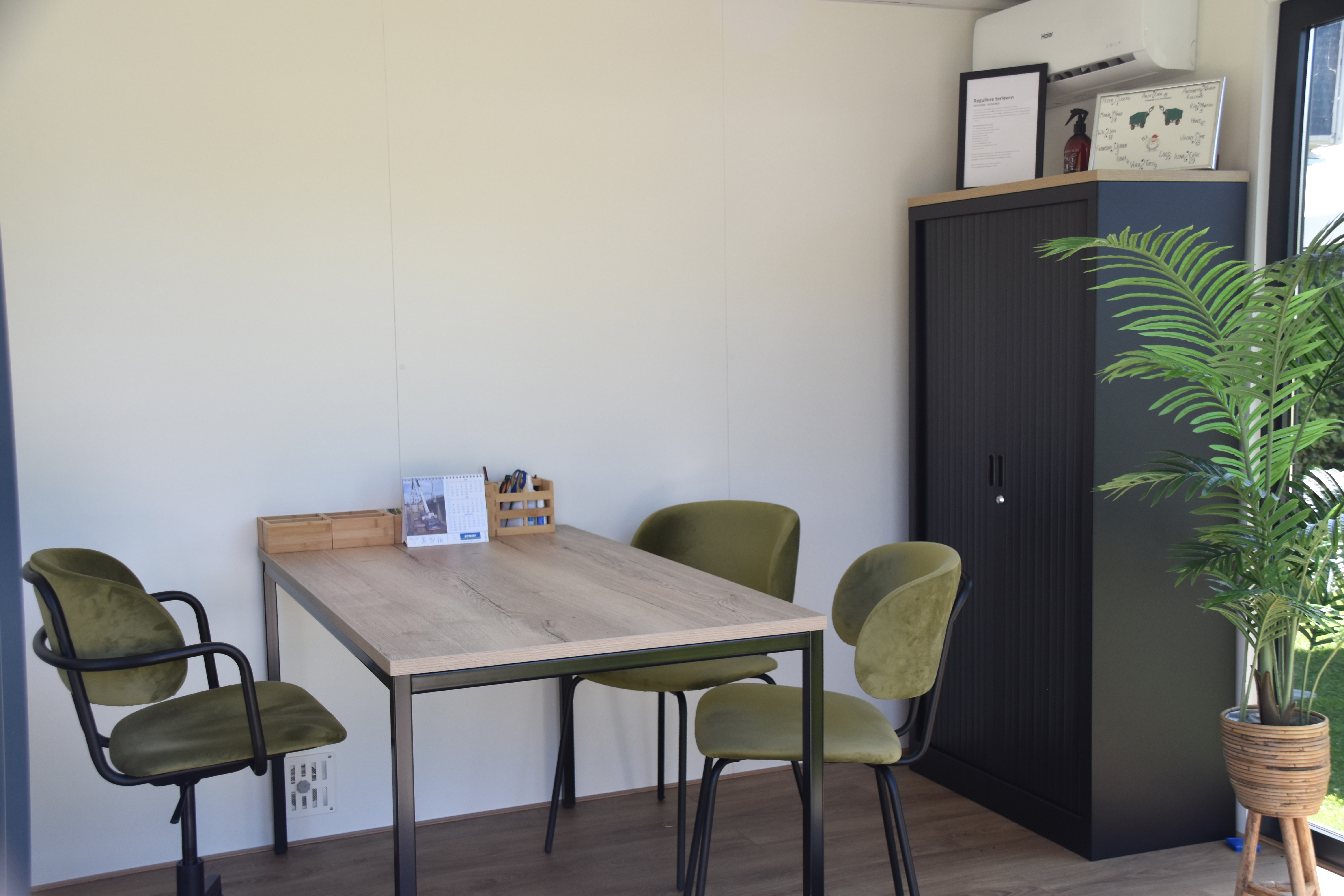 Camping-Büro mit Tisch und Stühlen, Schrank an der Wand und Topfpflanze im Vordergrund.