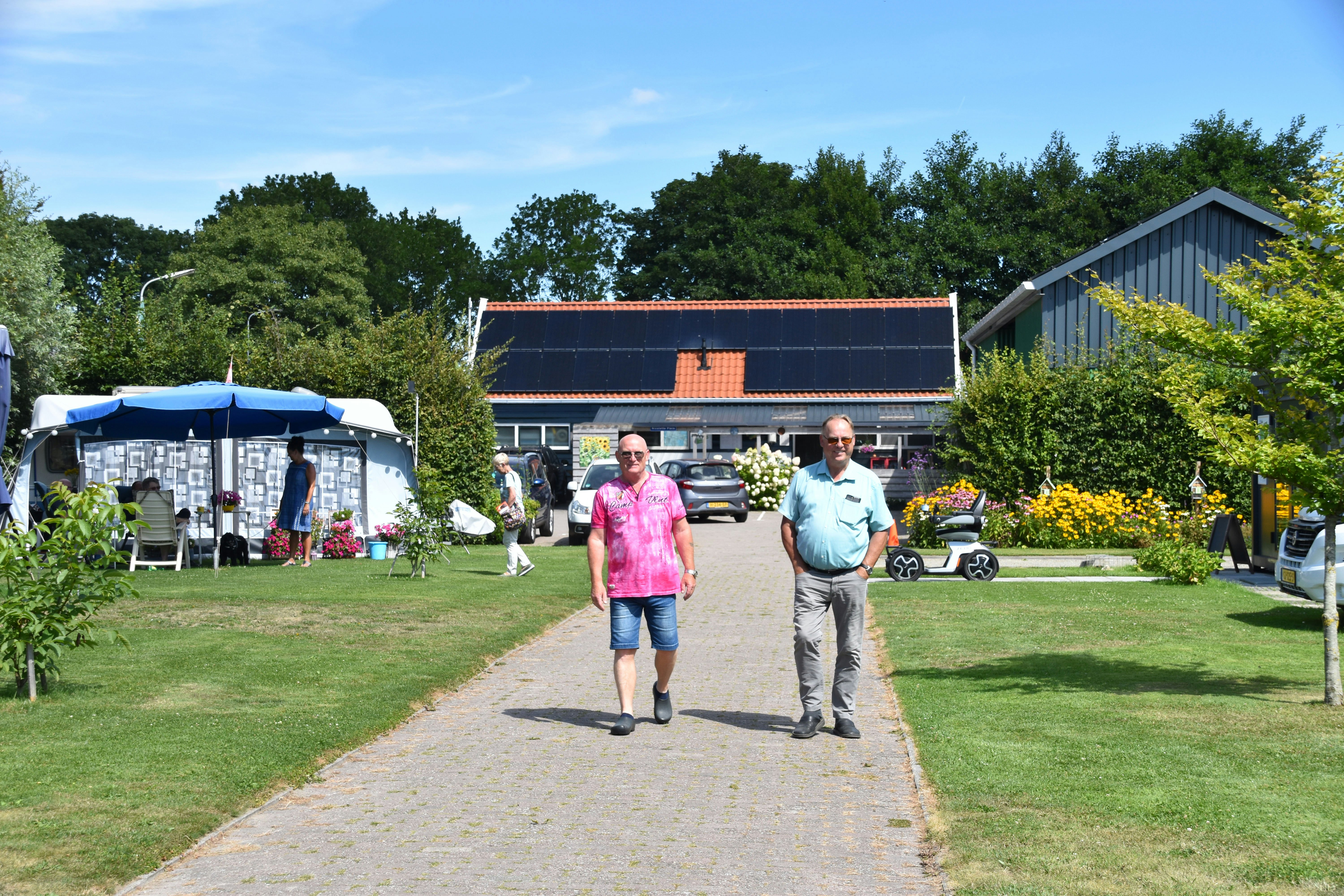 Zwei Männer gehen auf einem gepflasterten Weg am Campingplatz vorbei, im Hintergrund ein Gebäude und Zelte mit Gartenbereich.