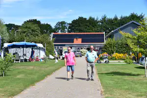 Zwei Männer gehen auf einem gepflasterten Weg am Campingplatz vorbei, im Hintergrund ein Gebäude und Zelte mit Gartenbereich.