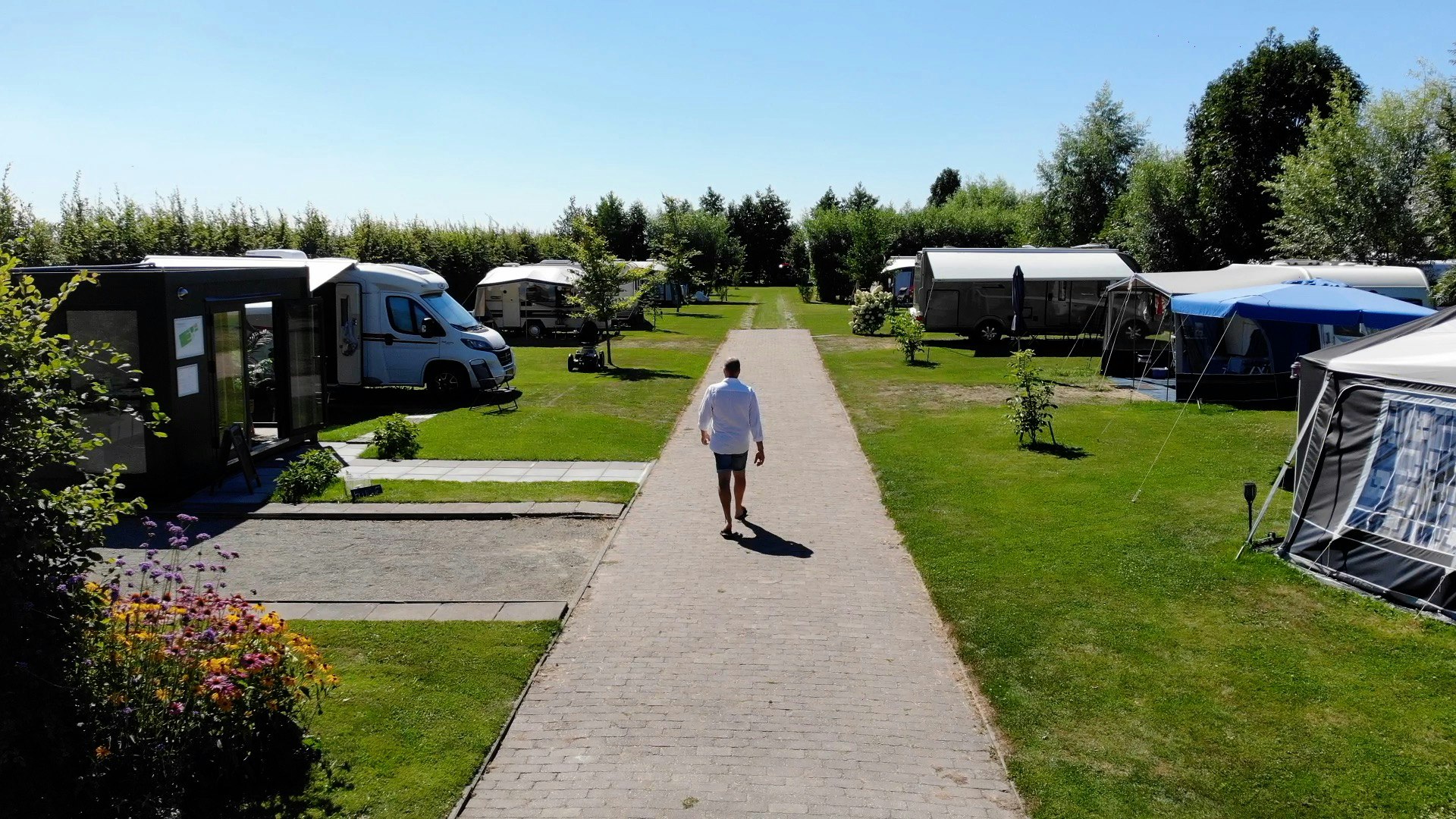 Campingplatz-Gasse mit Wohnmobilen und Wohnwagen, Rasenflächen und Hecken, im Hintergrund Bäume und ein Weg zur Rezeption.