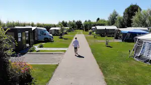 Campingplatz-Gasse mit Wohnmobilen und Wohnwagen, Rasenflächen und Hecken, im Hintergrund Bäume und ein Weg zur Rezeption.
