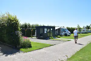 Wohnmobil und Wohnwagen auf dem Campingplatz, Weg und Rasen mit Blumenbeeten vor einem kleinen Gartenhäuschen.