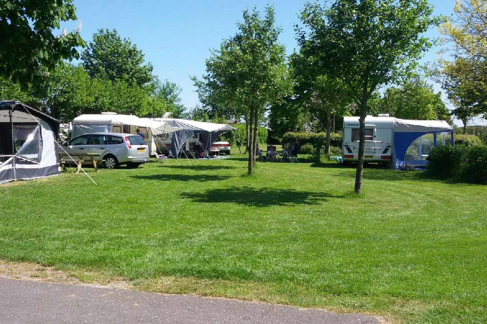 Campingplatz mit Rasenfläche, mehreren Wohnwagen und Zelten zwischen Bäumen, im Hintergrund ein Parkplatz mit Fahrzeugen.