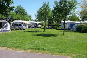 Campingplatz mit Rasenfläche, mehreren Wohnwagen und Zelten zwischen Bäumen, im Hintergrund ein Parkplatz mit Fahrzeugen.
