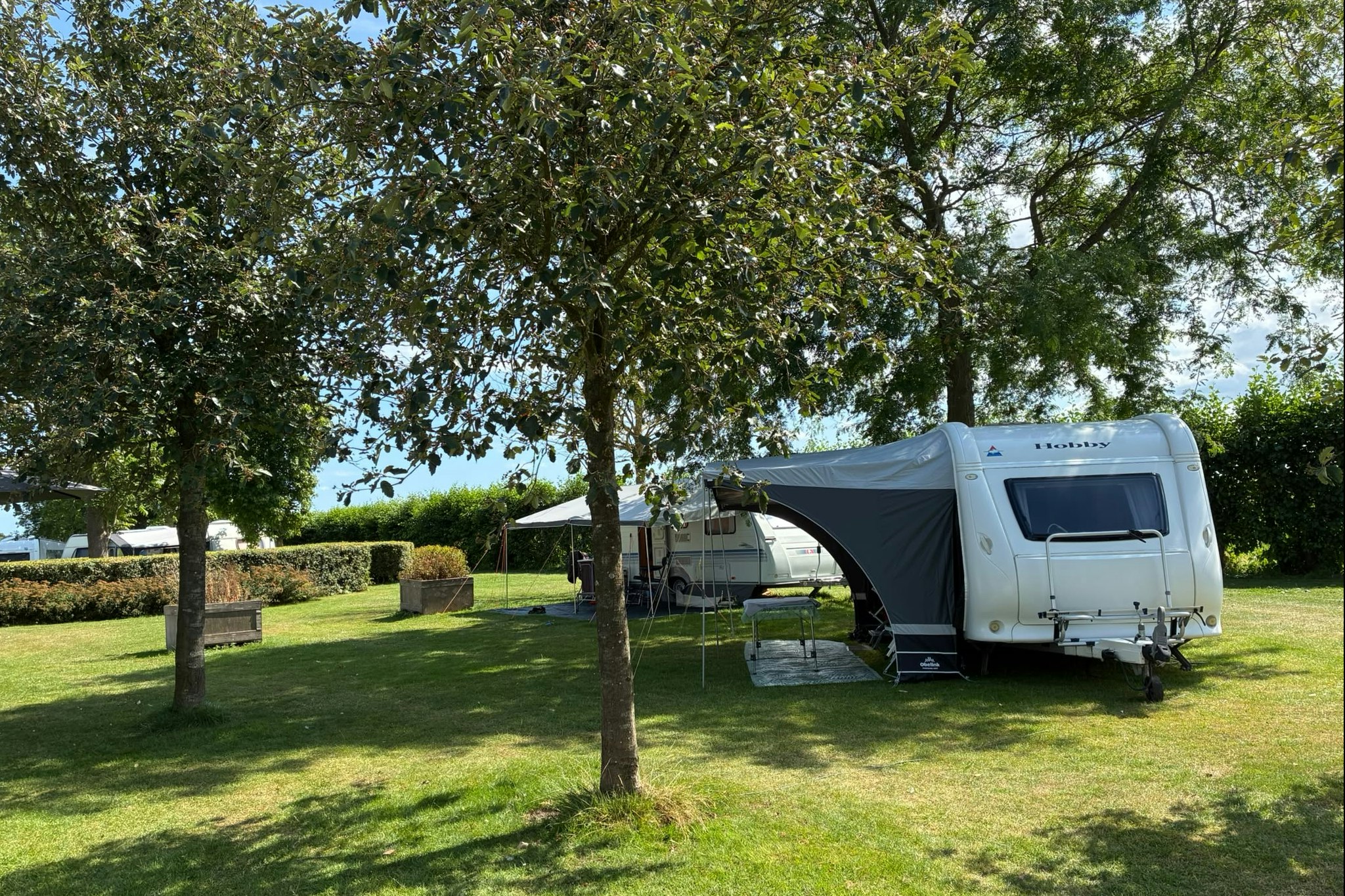 Wohnwagen mit Vorzelt auf einem Campingplatz, umgeben von Rasen, Hecken und Bäumen.