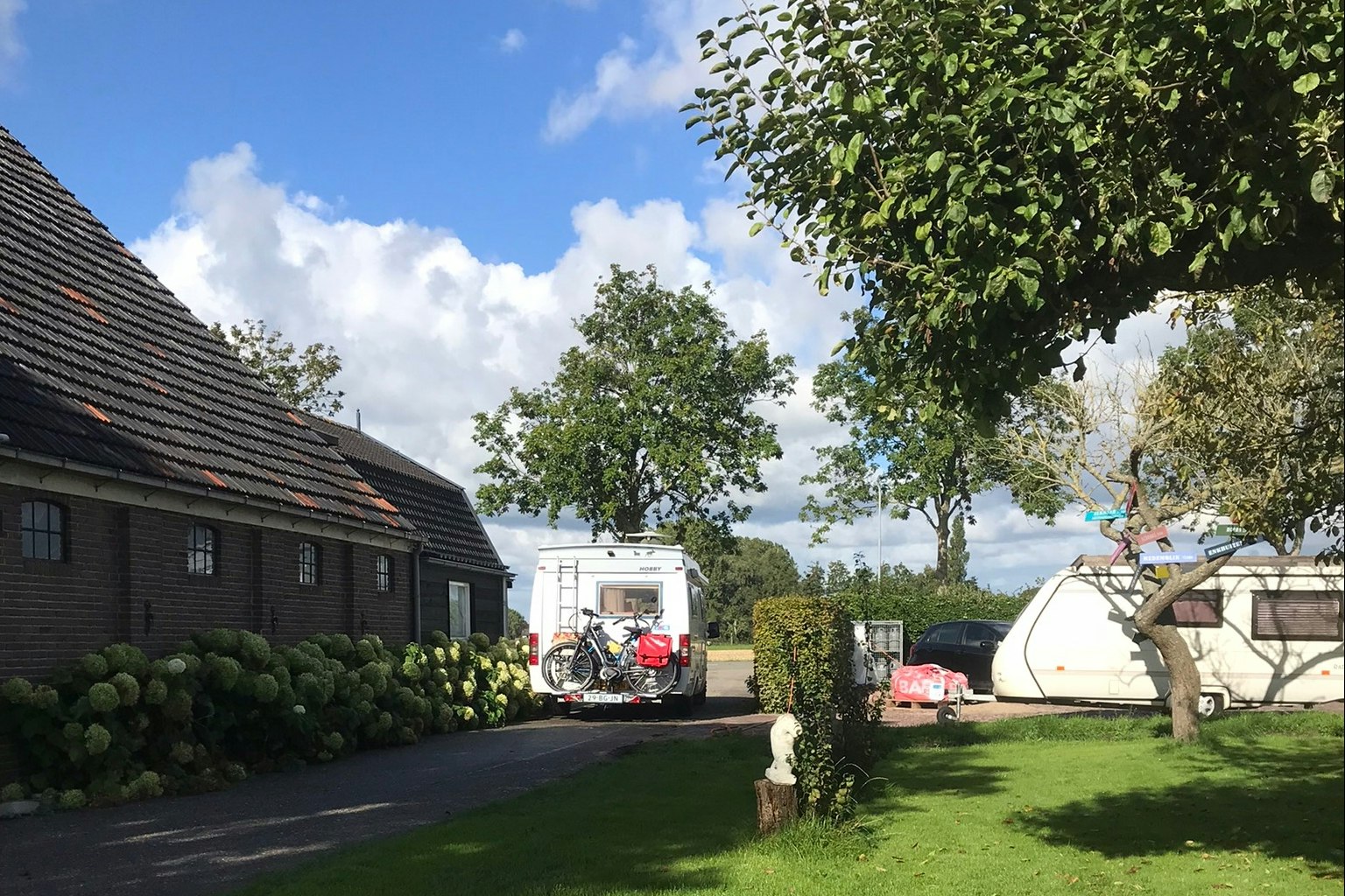 Wohnwagen und Wohnmobil auf einem Campingplatz, gepflasterter Weg und Rasen mit Bäumen vor kleinen Gebäuden.