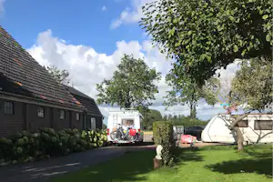 Wohnwagen und Wohnmobil auf einem Campingplatz, gepflasterter Weg und Rasen mit Bäumen vor kleinen Gebäuden.