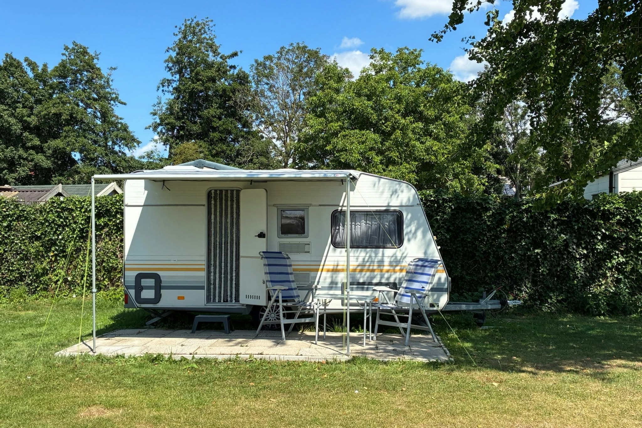 Wohnwagen mit Vorzelt auf einem Stellplatz im Grünen, mit Tisch und Stühlen vor dem Eingang, umgeben von Bäumen.