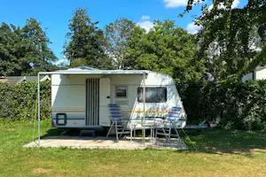 Wohnwagen mit Vorzelt auf einem Stellplatz im Grünen, mit Tisch und Stühlen vor dem Eingang, umgeben von Bäumen.