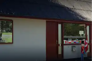 Eingang eines Campinghauses mit brauner Tür, Wand, Flaggen und Infotafel vor dem Gebäude.