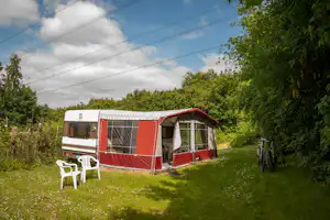 Wohnwagen mit rotem Vorzelt und großen Fenstern auf einer Wiese, daneben zwei weiße Campingstühle und ein Fahrrad.