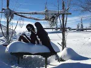 Schwarze Camping-Figur auf einer Bank, umgeben von Schnee, mit Bäumen und Zaun im Hintergrund.