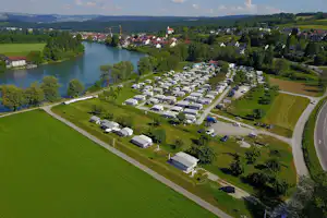 Luftaufnahme eines Campingplatzes mit Wohnwagen und Mobilheimen am Fluss, umgeben von Wiesen, Bäumen und einem Dorf.