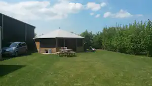 Zelt und Sitzgruppe mit Tisch auf dem Rasen, daneben ein Wohnwagen und ein Schuppen, Hecken und Bäume im Hintergrund.