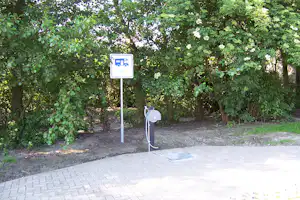 Wasserstelle am Campingplatz mit Zapfsäule und Schildern, umgeben von Bäumen und Sträuchern.