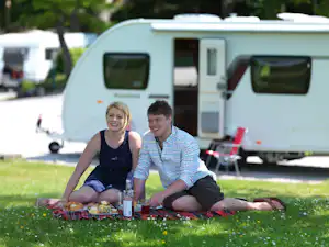 Ein Paar sitzt beim Picknick auf einer Decke im Grünen vor einem Wohnwagen auf dem Campingplatz.