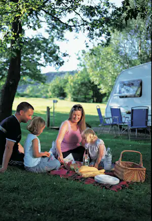 Picknick am Campingplatz im Grünen: Familie sitzt auf einer Decke, Essen und Getränke, Campingstühle und Auto im Hintergrund.