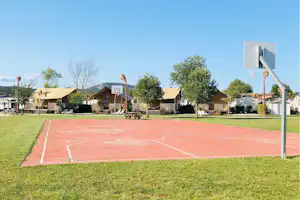 Basketballfeld auf einem Campingplatz mit rotem Spielfeld, weißen Linien und mehreren Bungalows im Hintergrund, umgeben von Rasen und Bäumen.