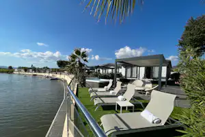 Terrasse am See mit mehreren Sonnenliegen, Sonnenschirmen und Blick auf das Wasser, daneben ein Steg und Gebäude.