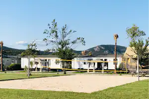 Campinganlage mit Wohnwagen und überdachten Terrassen, Rasenfläche, Bäumen und Wegen vor weißen Gebäuden.