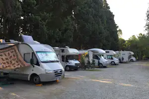 Reihe von Wohnmobilen auf einem Campingplatz, Kiesfläche und hohe Bäume im Hintergrund.