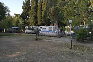Wohnwagen auf einem Campingplatz, umgeben von Bäumen, mit Laterne und Rasenfläche im Vordergrund.